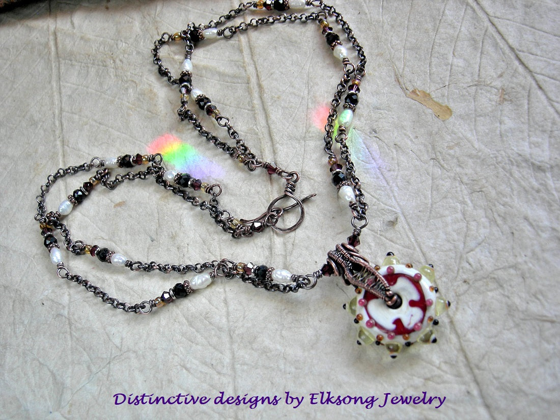 Copper chain necklace with a lampwork glass focal on a textured surface, featuring 'Distinctive designs by Elksong Jewelry'.