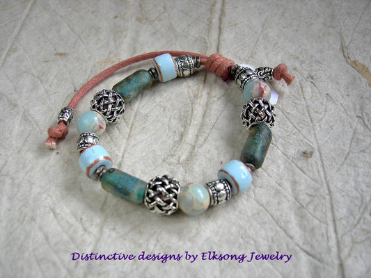 Bracelet with silver & blue glass, ceramic & stone beads on cord and featuring 'Distinctive designs by Elksong Jewelry'.